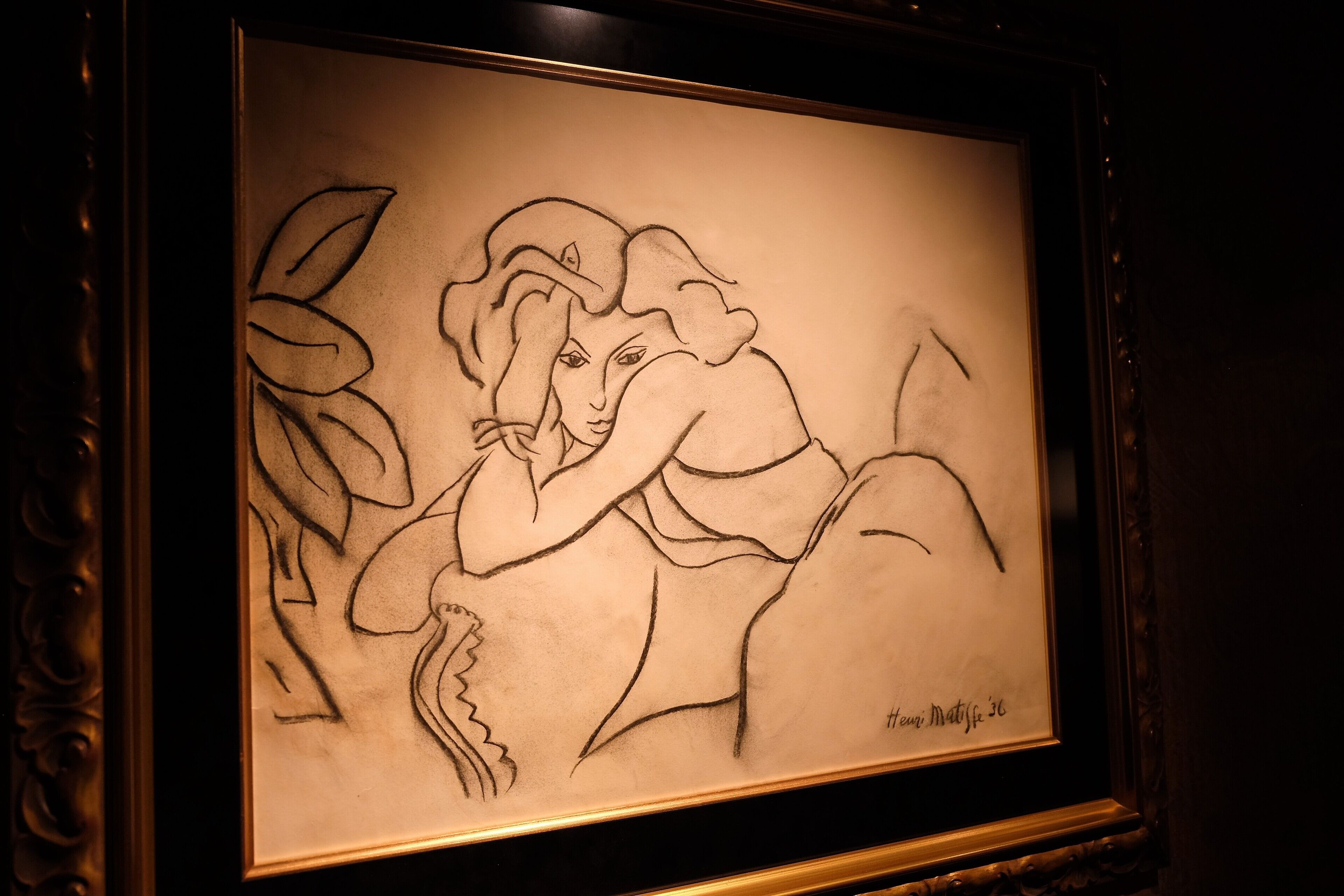 Une œuvre d’art encadrée, portant une signature, est exposée dans une pièce sombre dont les bords sont ornés de cadres dorés.