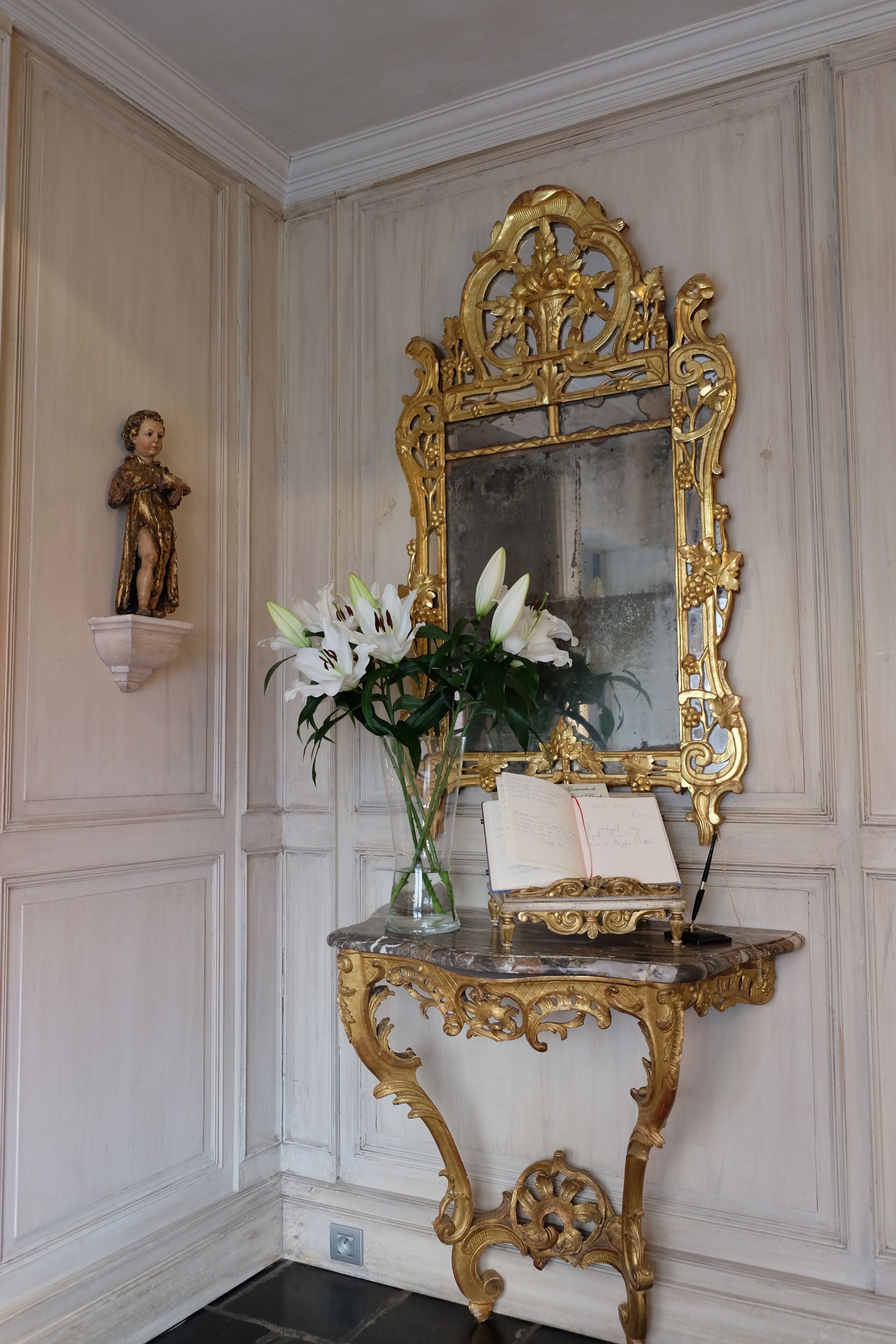 A room with a gold-framed mirror, a white marble console table with a book and flowers, and a small statue on the wall.