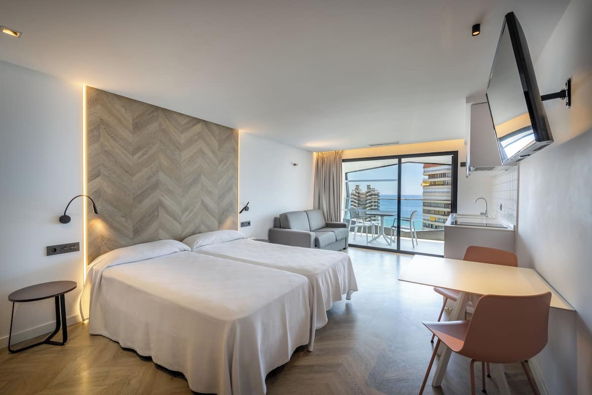 A modern hotel room with two beds, a dining area, and a view of the sea.
