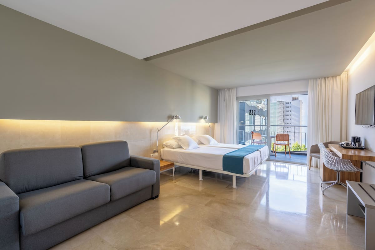 A modern hotel room with a sofa, a bed, a desk, and a balcony with a view of the city.