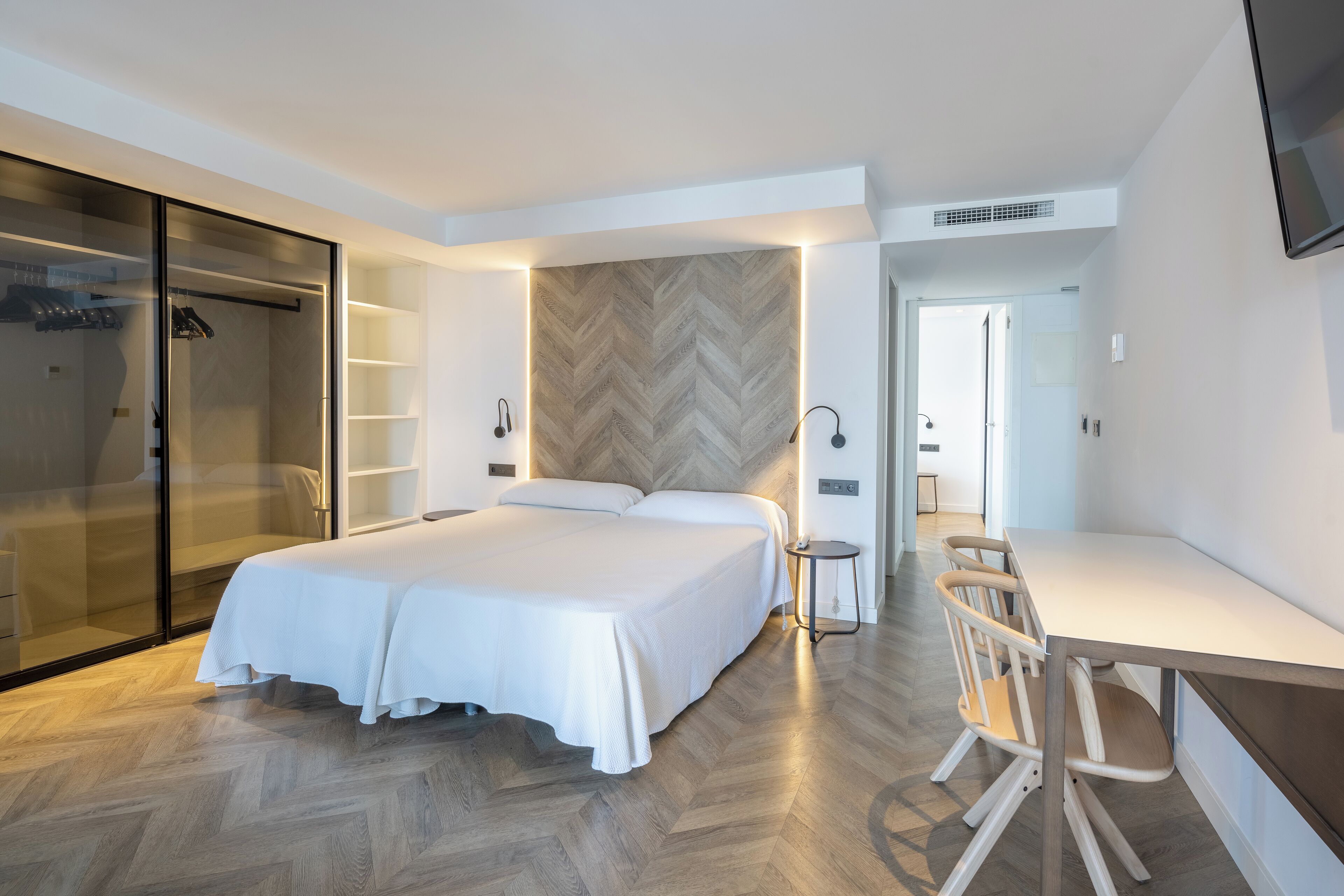 Un dormitorio moderno con una cama grande, un escritorio y una silla. Hay un armario con puertas correderas y una pared con patrón de espiga.