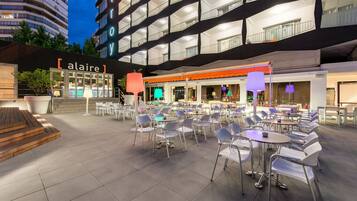 Un área de café al aire libre con varias mesas y sillas, un edificio con balcones y un letrero que dice "alair".