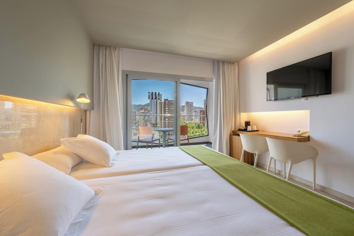 A hotel room with two beds, a desk, a chair, a TV, and a balcony with a city view.