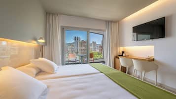 Ein Hotelzimmer mit zwei Betten, einem Schreibtisch, einem Sessel, einem Fernseher und einem Balkon mit Blick auf die Stadt.