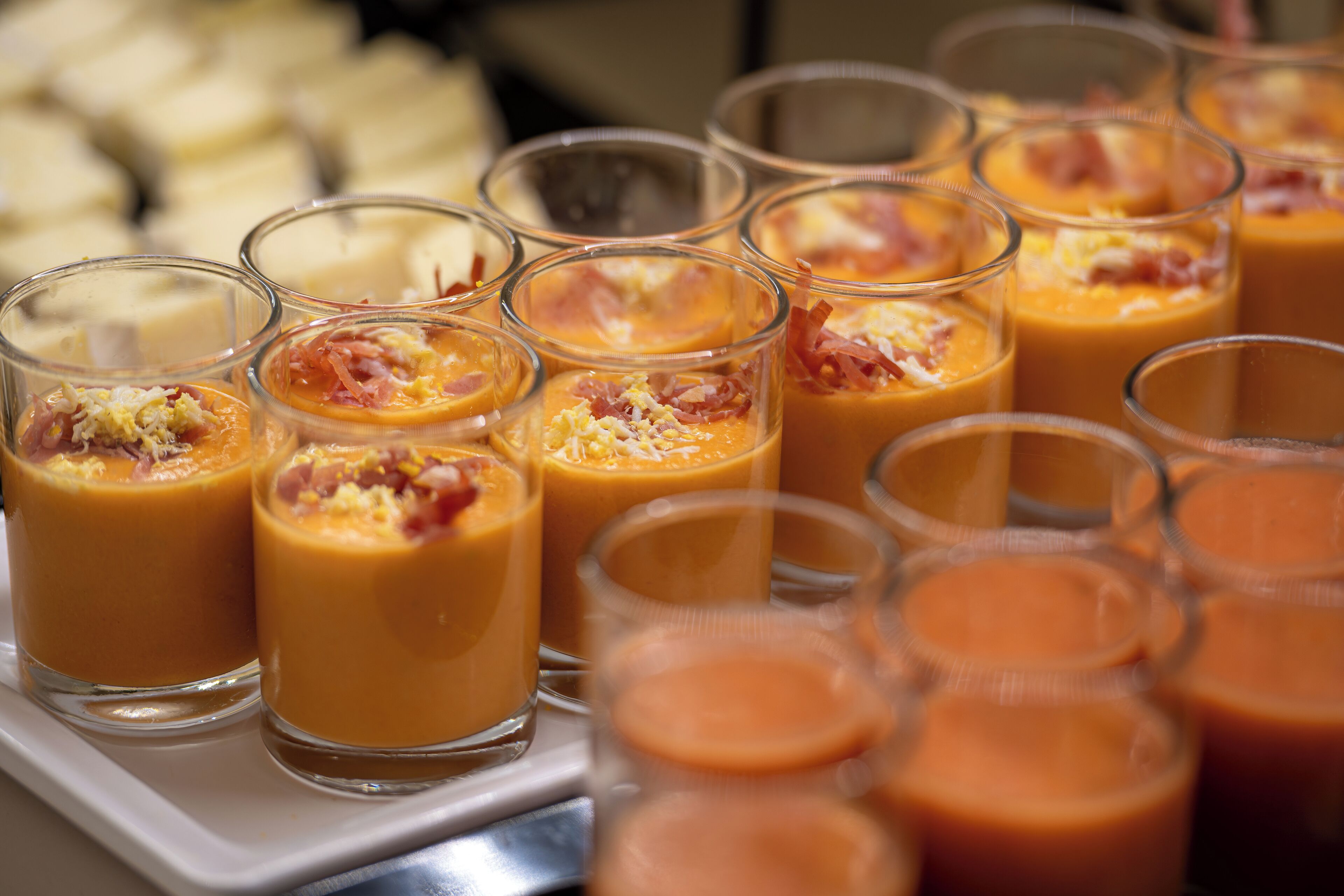 A tray of small glasses filled with a creamy orange beverage, garnished with bits of bacon and cheese, alongside a variety of other food items.