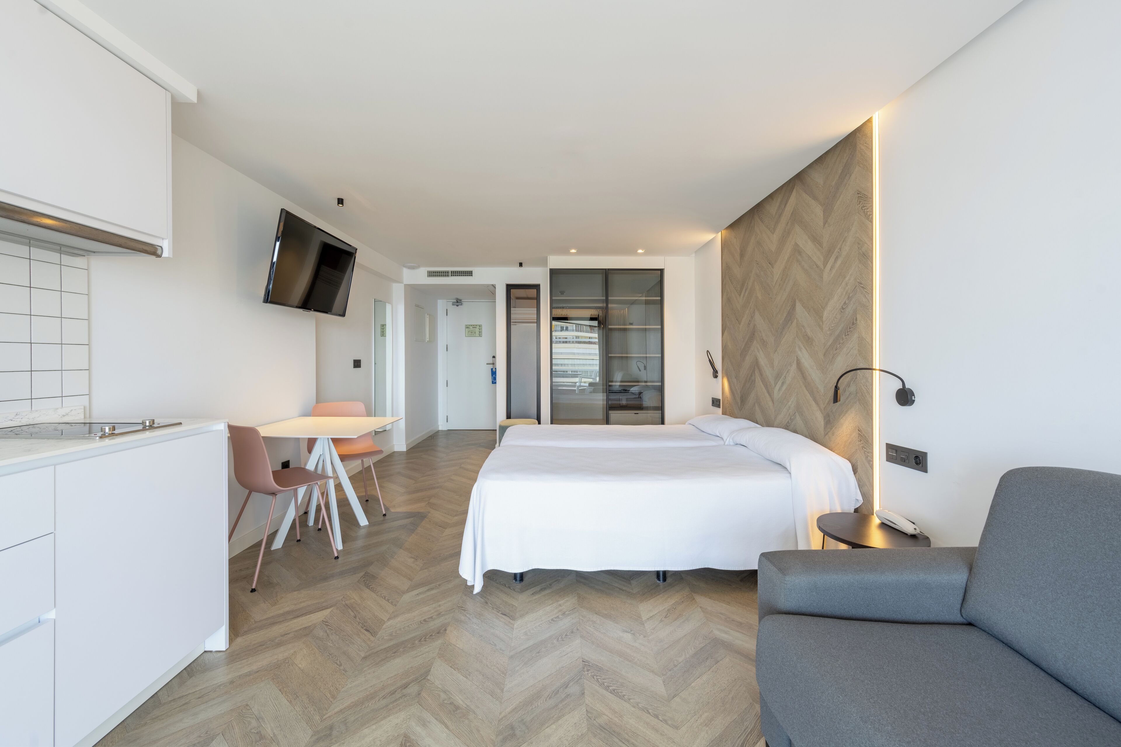 A modern hotel room with a bed, a sofa, a dining area with a table and chairs, a TV mounted on the wall, and a wooden herringbone floor.