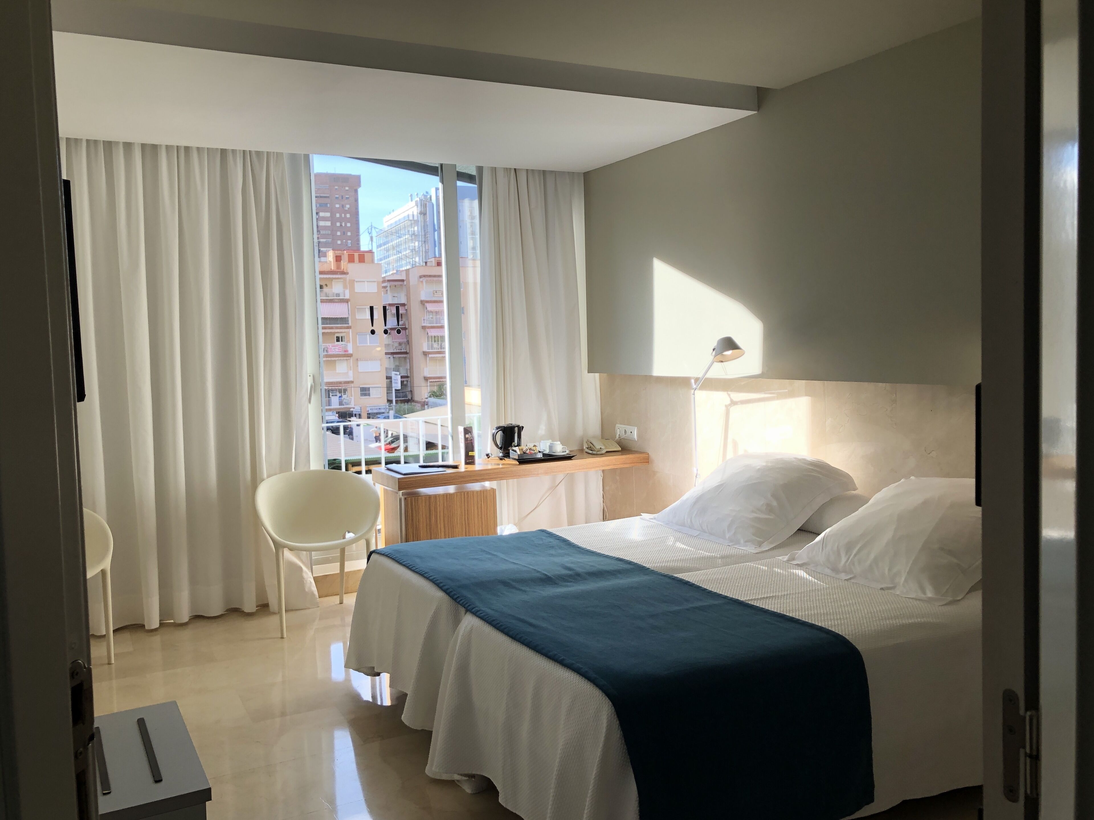 Habitación de hotel con cama, escritorio, silla y vista de la ciudad a través de amplios ventanales.