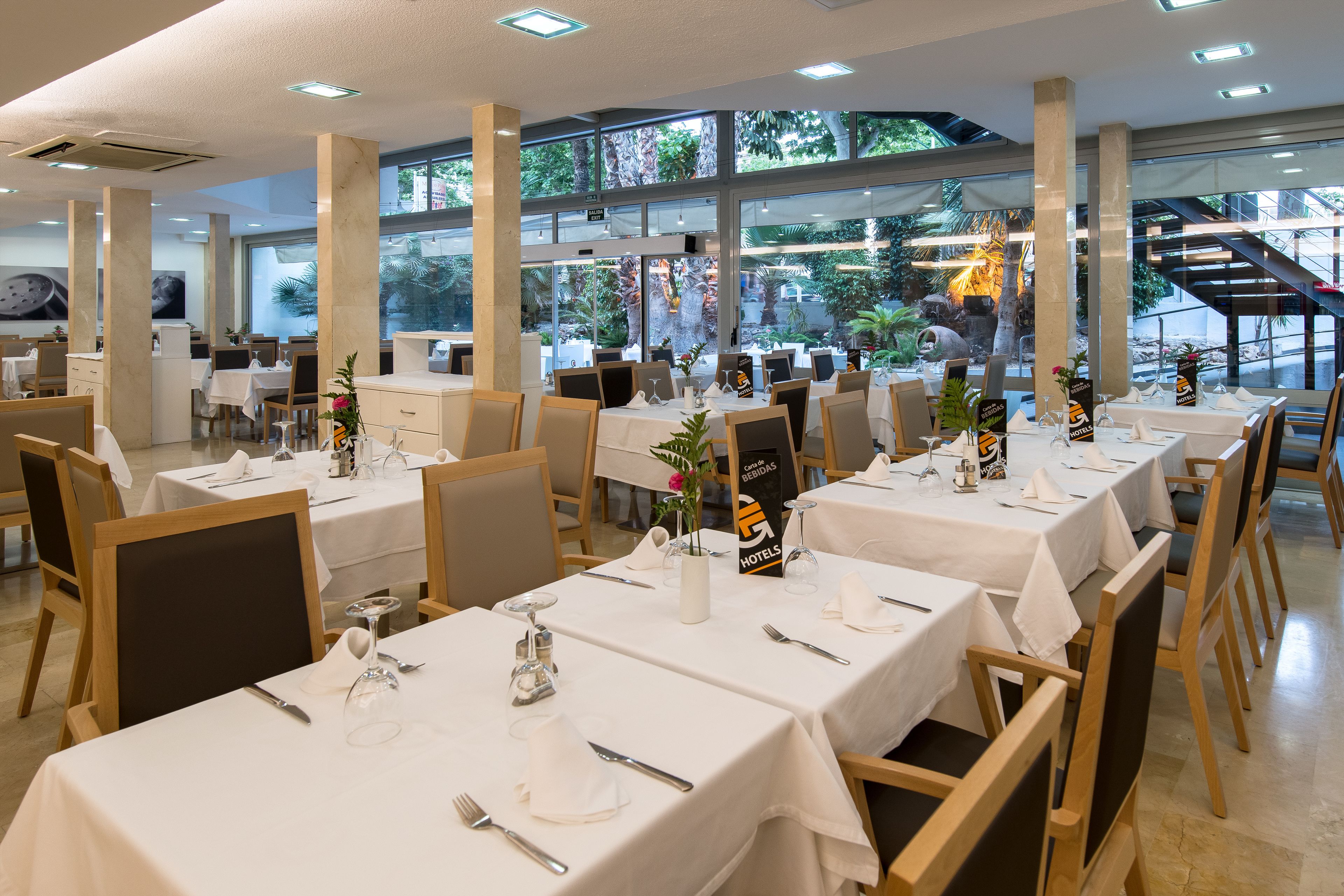 A restaurant with multiple tables set for dining, large windows, and a view of greenery outside.
