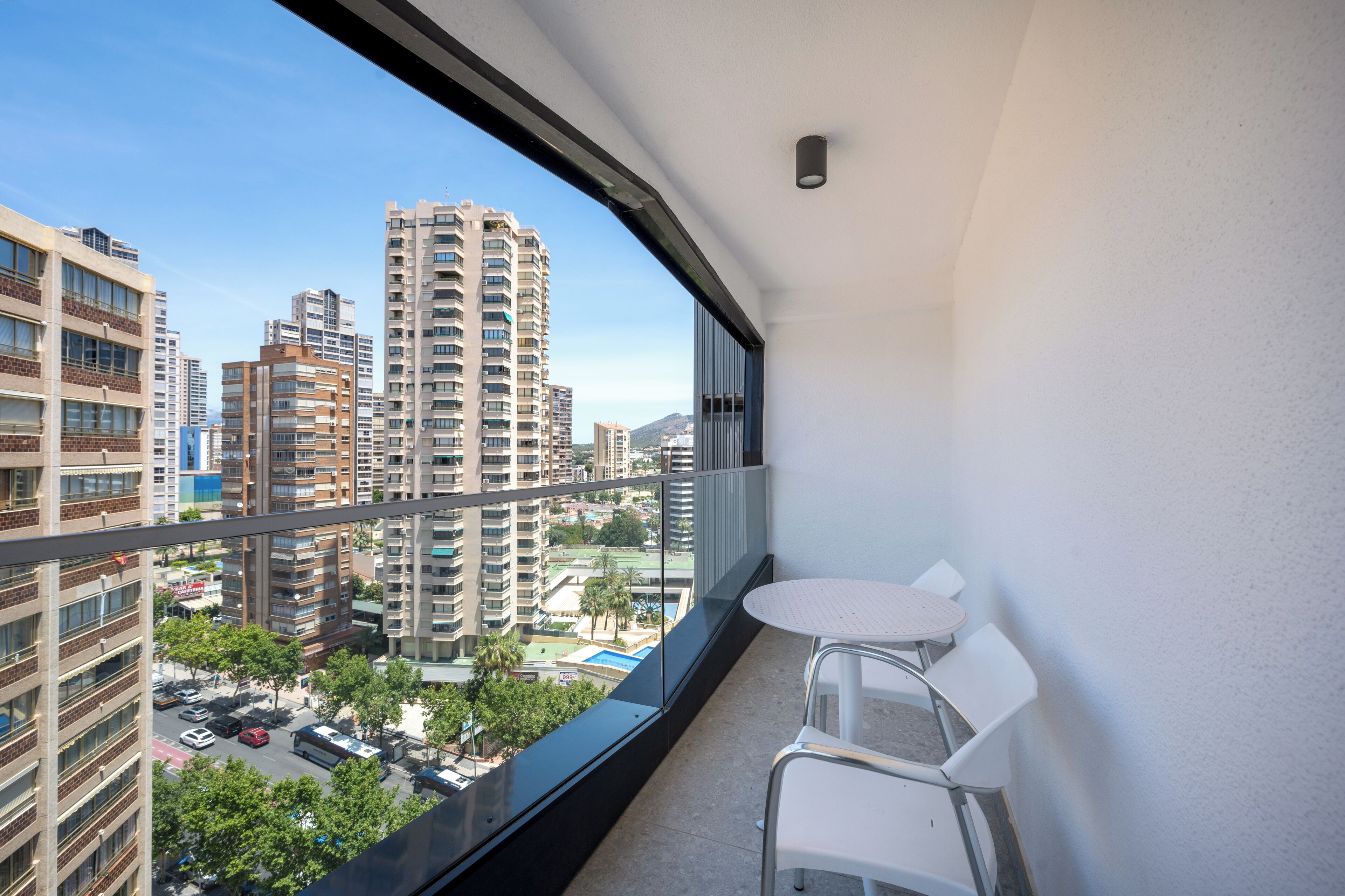 A balcony with a table and chairs, overlooking a cityscape with tall buildings and a clear sky.