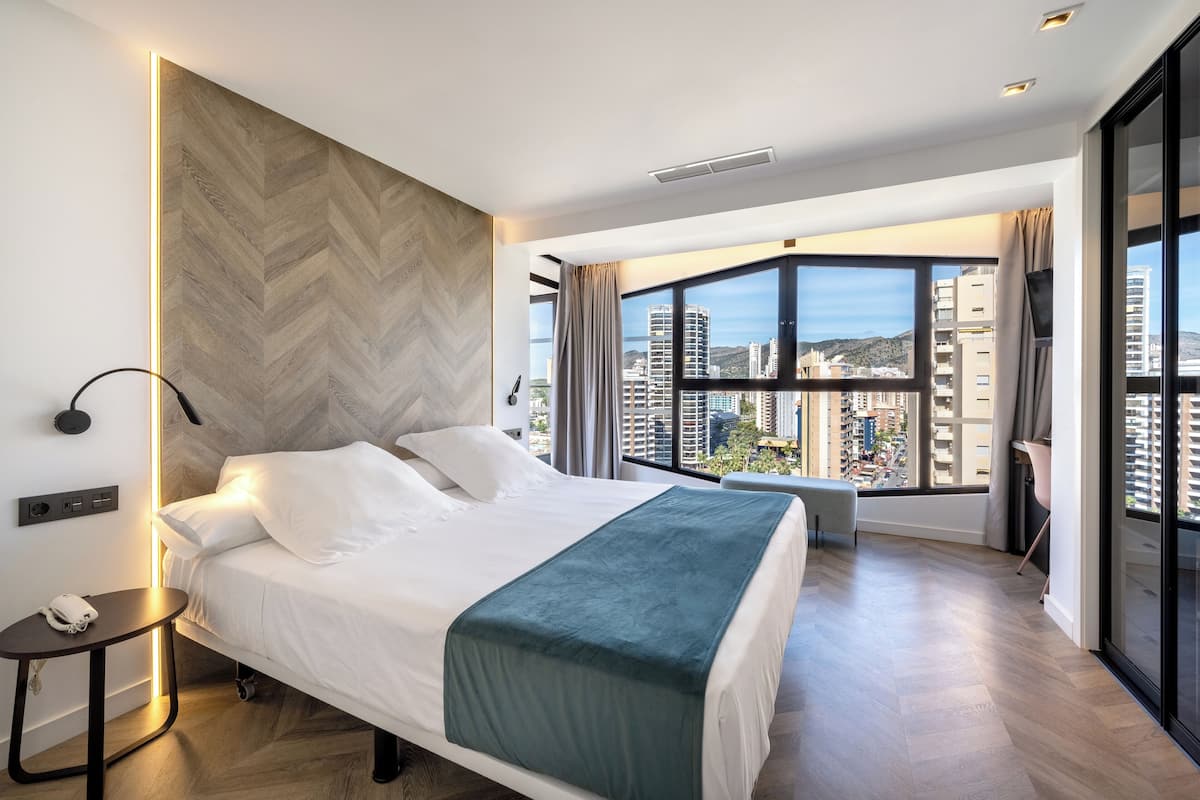 A modern hotel room with a large bed, a view of the city, and a herringbone-patterned wall.