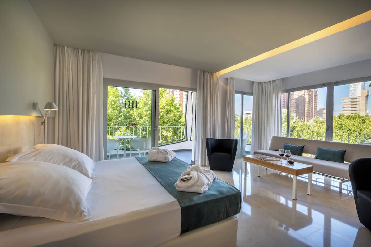 A modern hotel room with a large bed, a sofa, a dining table, and a balcony with a view of the city.