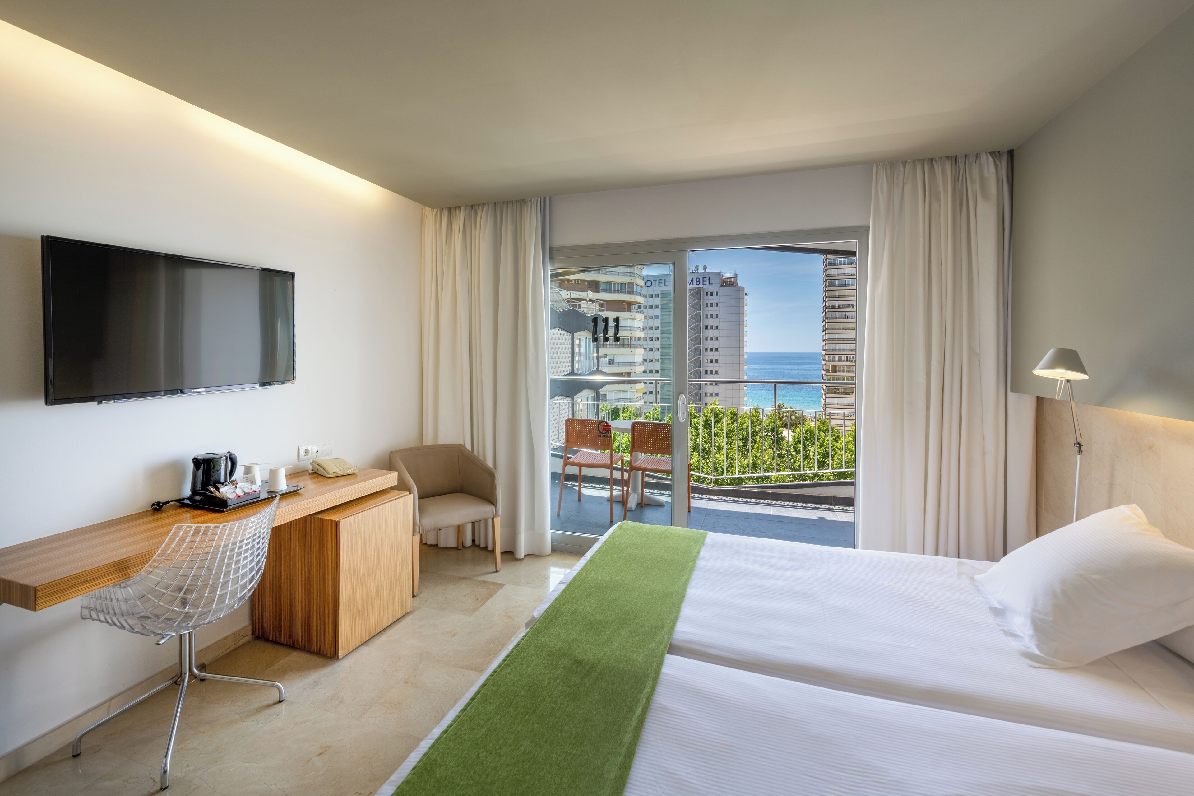 A hotel room with a bed, a desk with a chair, a TV, and a balcony with a view of the ocean and buildings.