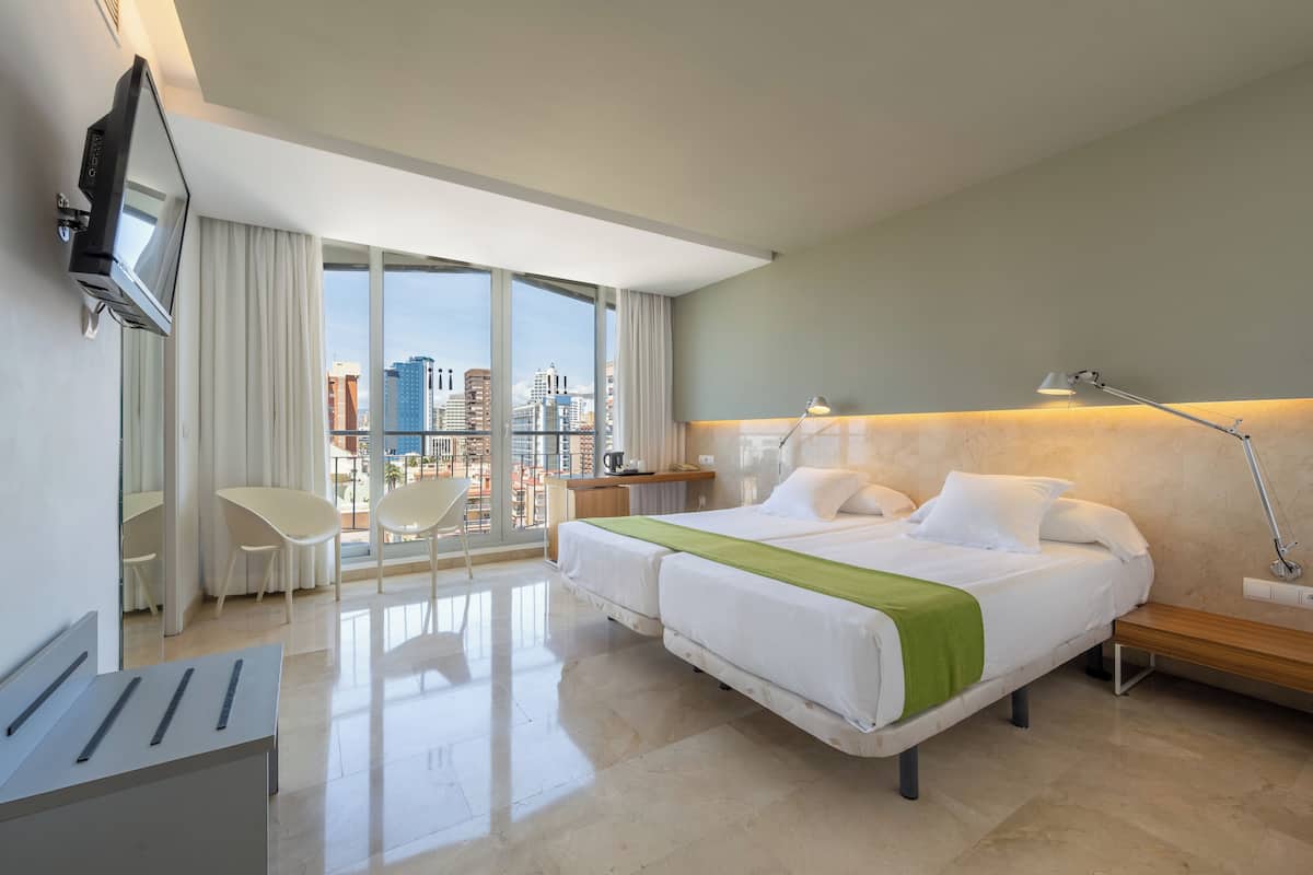 A hotel room with a bed, a desk, a chair, a TV, and a view of the city.