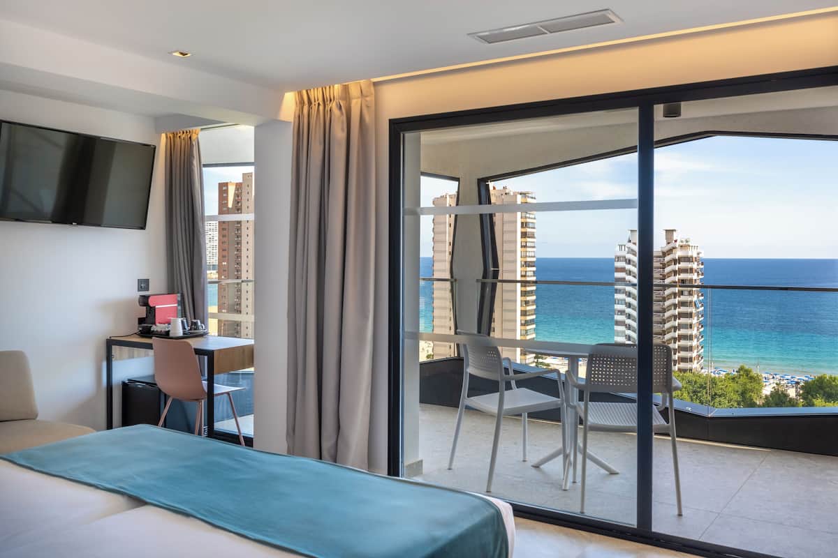A hotel room with a bed, a desk with a computer, a chair, and a balcony with a view of the ocean and buildings.