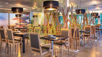 Un espace de restauration moderne avec des tables et des chaises en bois, des suspensions noires et des plantes d’intérieur.