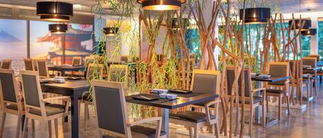 A modern dining area with wooden tables and chairs, black pendant lights, and indoor plants.