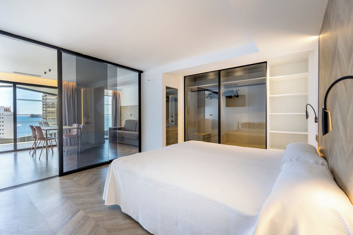 A modern hotel room with a large bed, a glass wall, and a view of the ocean.