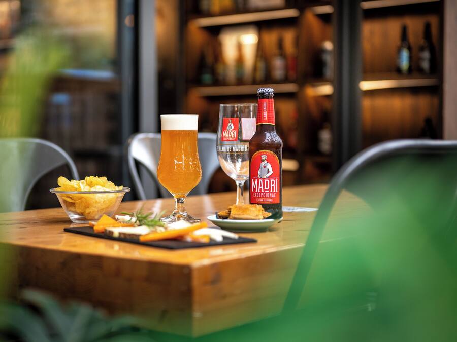 Une table avec un bol de chips, un verre de bière, une bouteille de bière Madri et un verre d’eau.