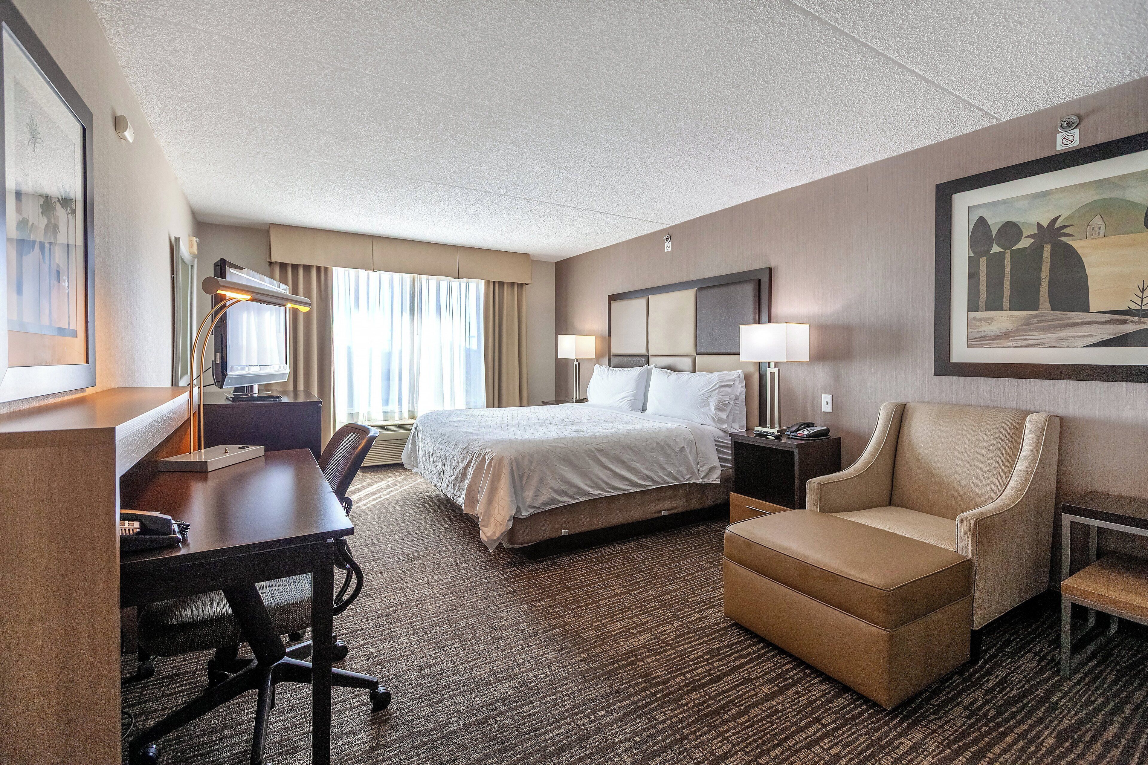 A hotel room with a bed, desk, chair, and a painting on the wall.