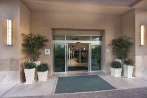Entrada de hotel con puerta de cristal, plantas en macetas y felpudo verde.