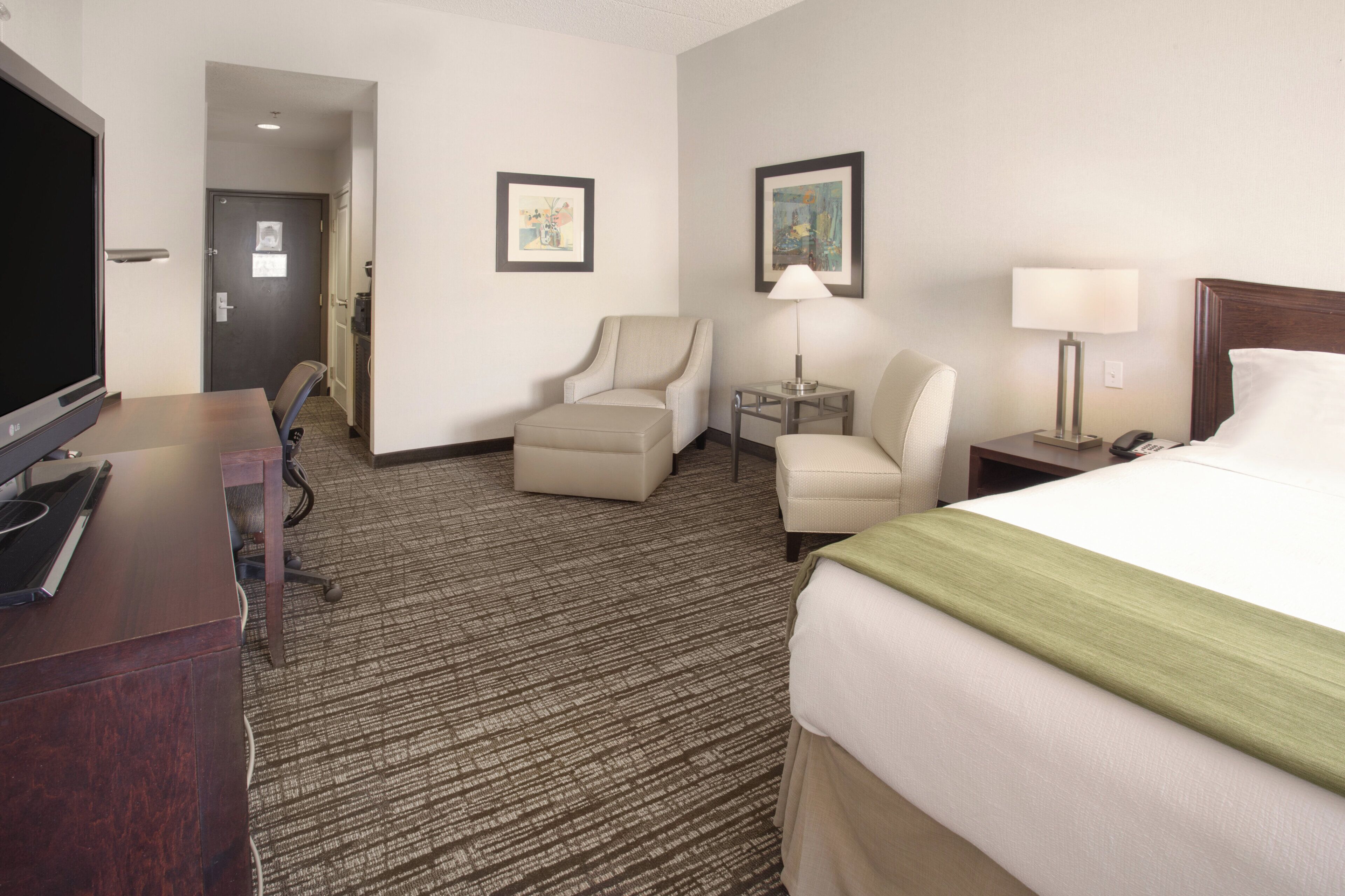 Habitación de hotel con cama, escritorio, dos sillas, televisor y obras de arte en las paredes.