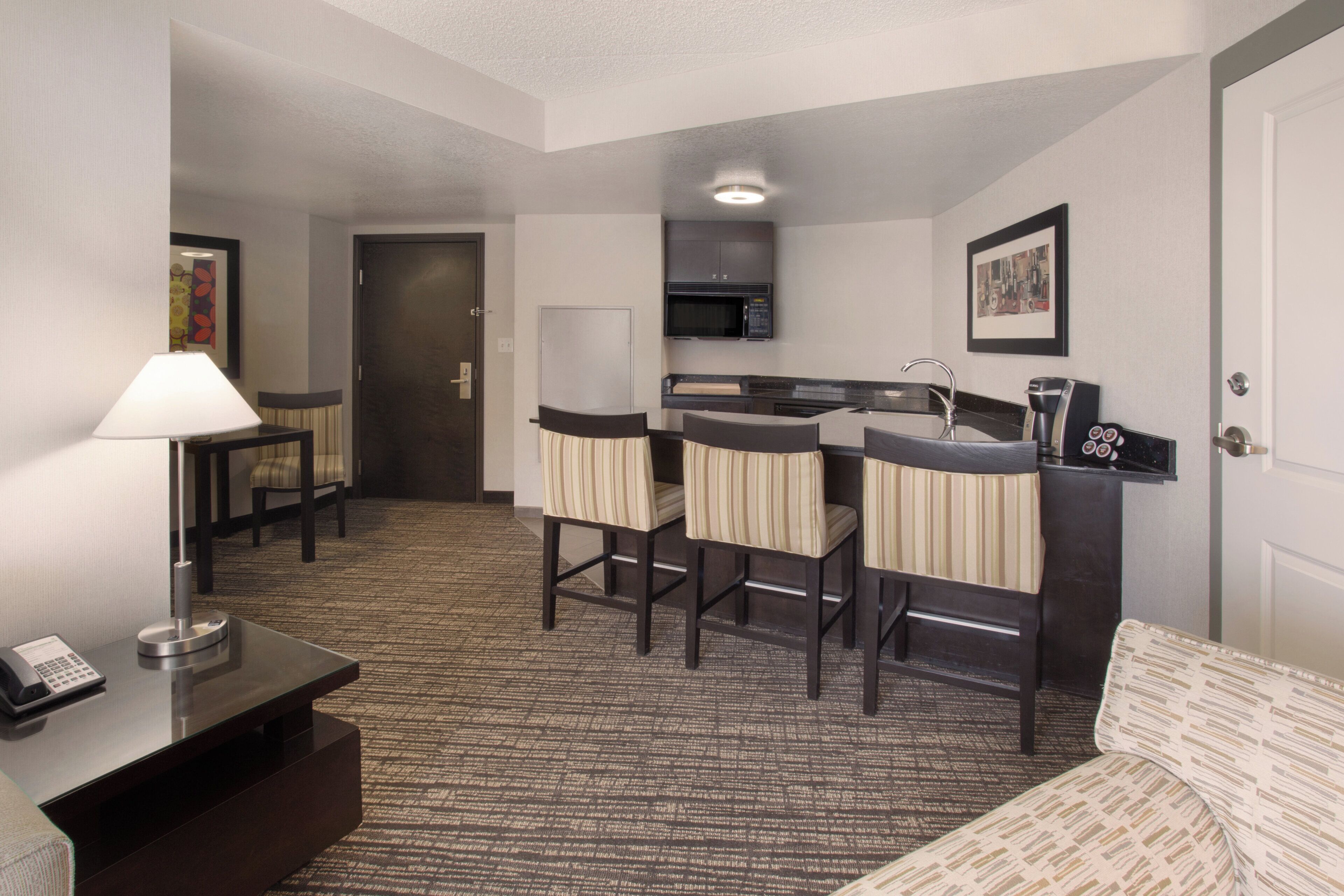 A hotel room with a kitchenette, a desk with a lamp, a sofa, and a chair.