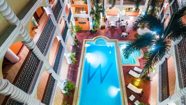 Ovenudblik over en hotelgård med en central swimmingpool, omgivet af palmer og udendørs siddeområder.