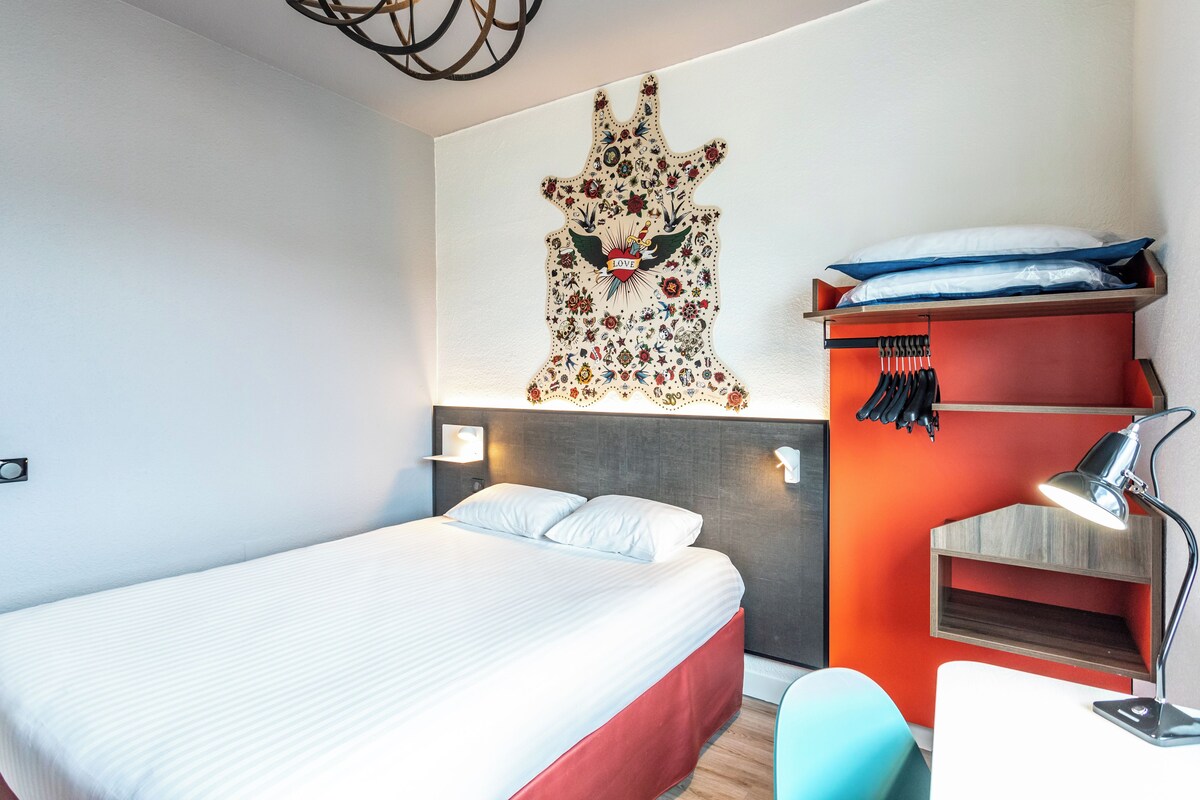 Una habitación de hotel moderna con una cama grande, un escritorio con lámpara, un estante en la pared con bolsos y un adorno decorativo en la pared.