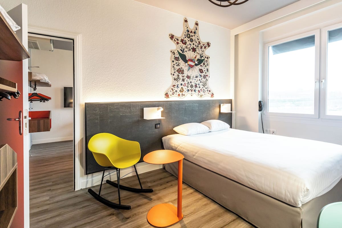A hotel room with a bed, a small table, a yellow chair, and a wall-mounted artwork.