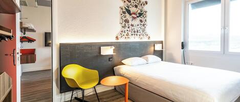 A hotel room with a bed, a small table, a yellow chair, and a wall-mounted artwork.