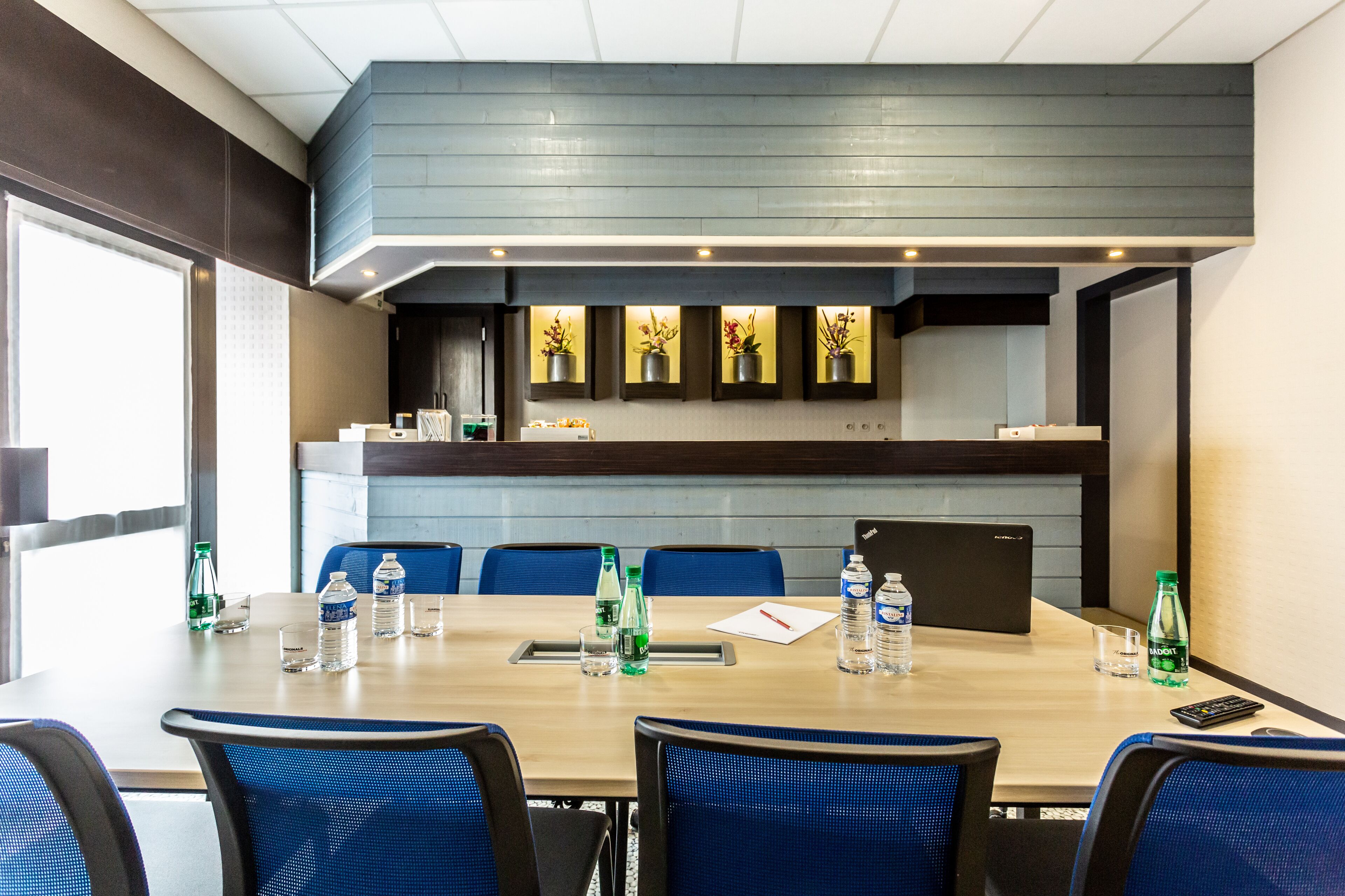 A conference room with a long table, blue chairs, water bottles, and a remote control.