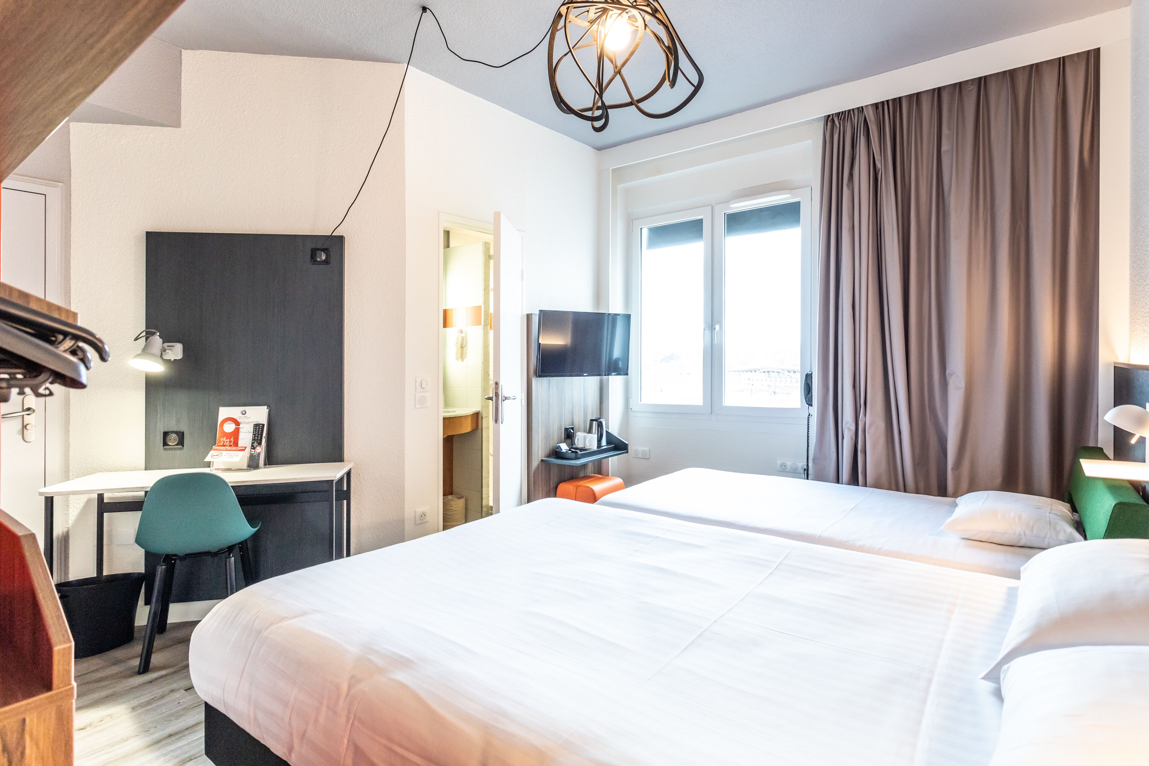 A hotel room with two beds, a desk, a chair, a TV, and a window with curtains.