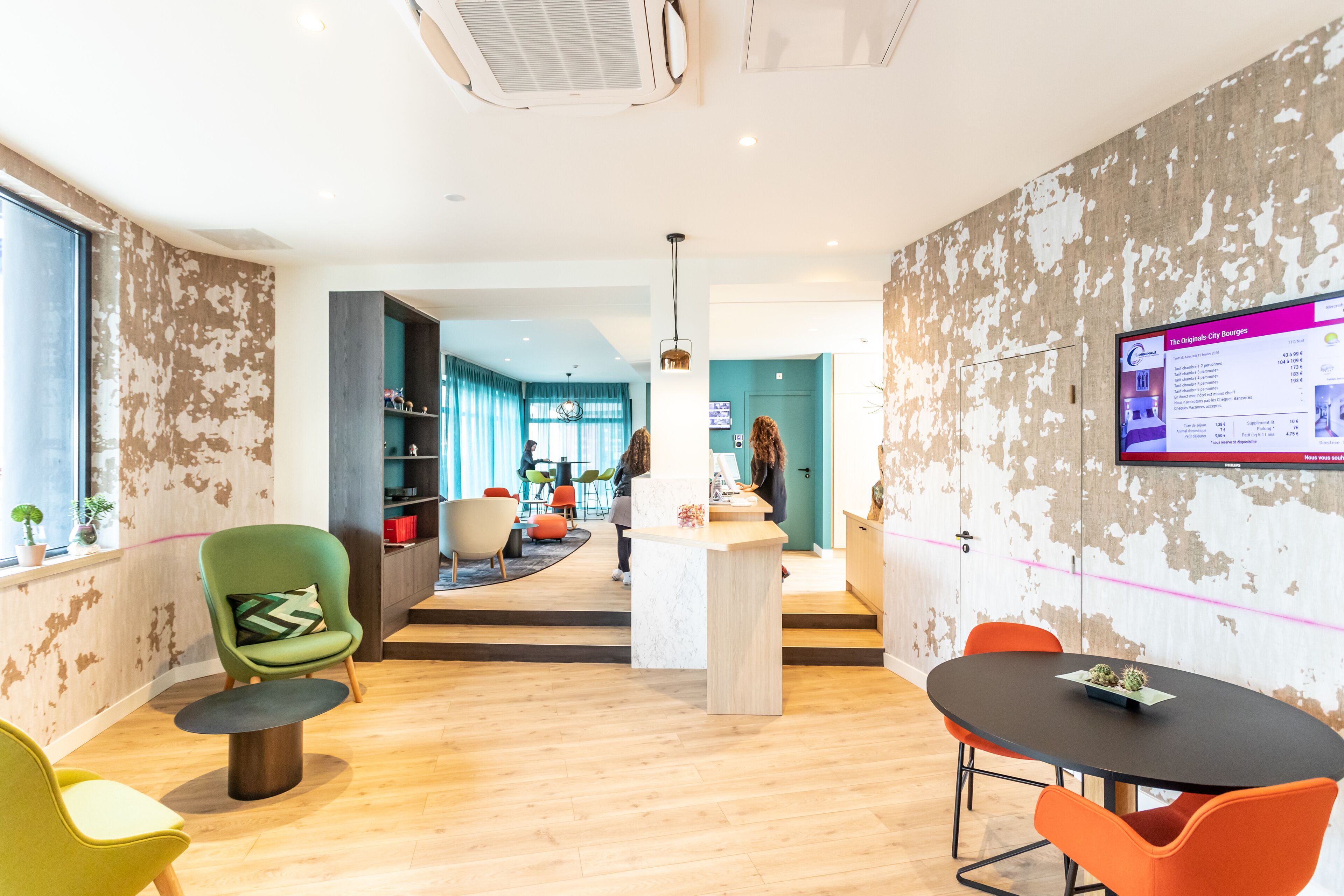 A modern office space with a reception desk, a large screen displaying content, and a mix of seating areas including a green chair and an orange chair.