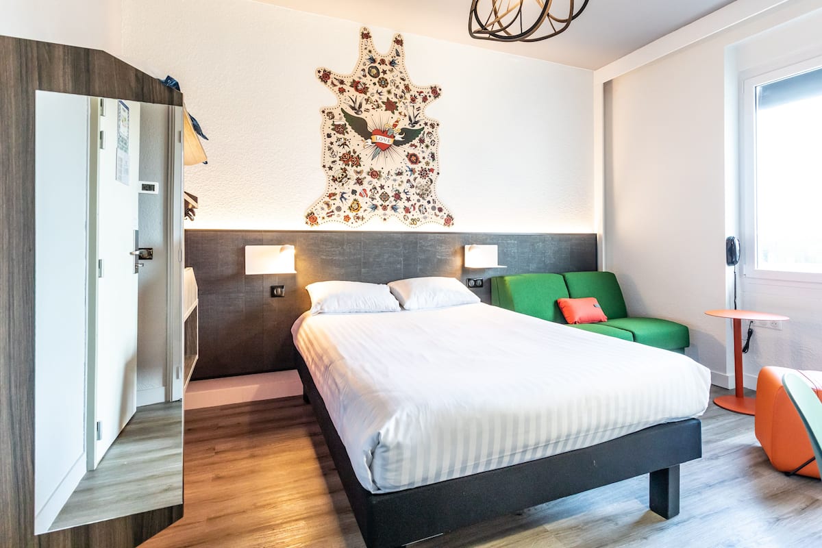 A modern hotel room with a large bed, a green sofa, a small orange table, and a wall-mounted artwork.
