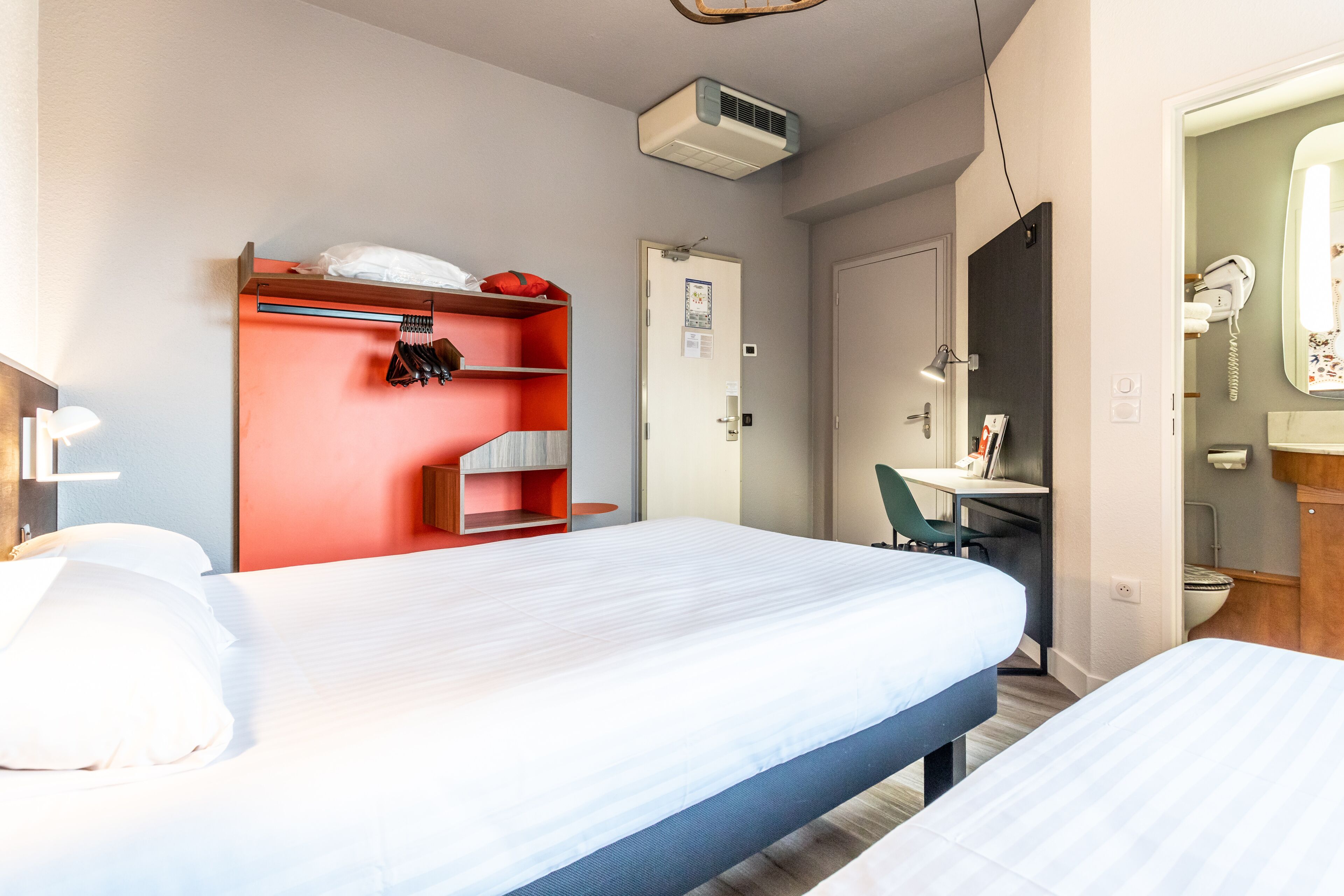 Habitación de hotel con cama, escritorio y silla, armario y baño con espejo e inodoro.