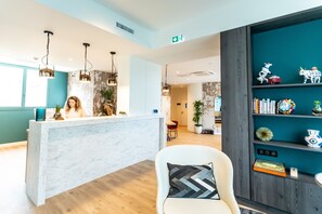 Un comptoir d’accueil avec un plateau en marbre, une chaise blanche avec un coussin à motifs et un mur de couleur turquoise orné d’étagères où sont exposés divers objets.