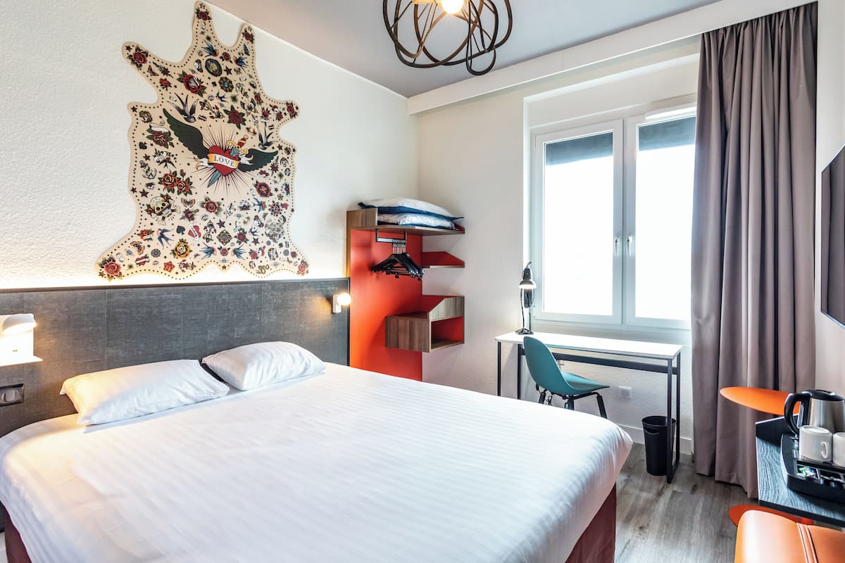 A hotel room with a bed, a desk, a chair, a coat rack, and a window with curtains.