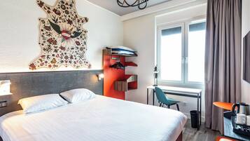 A hotel room with a bed, a desk, a chair, a coat rack, and a window with curtains.