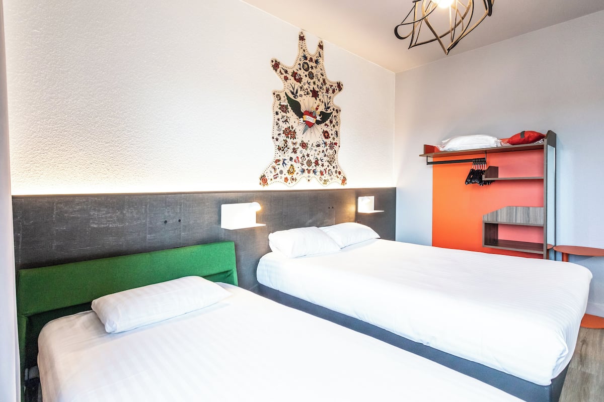 A hotel room with two beds, a green headboard, a wall-mounted artwork, and a small shelf with a red pillow.