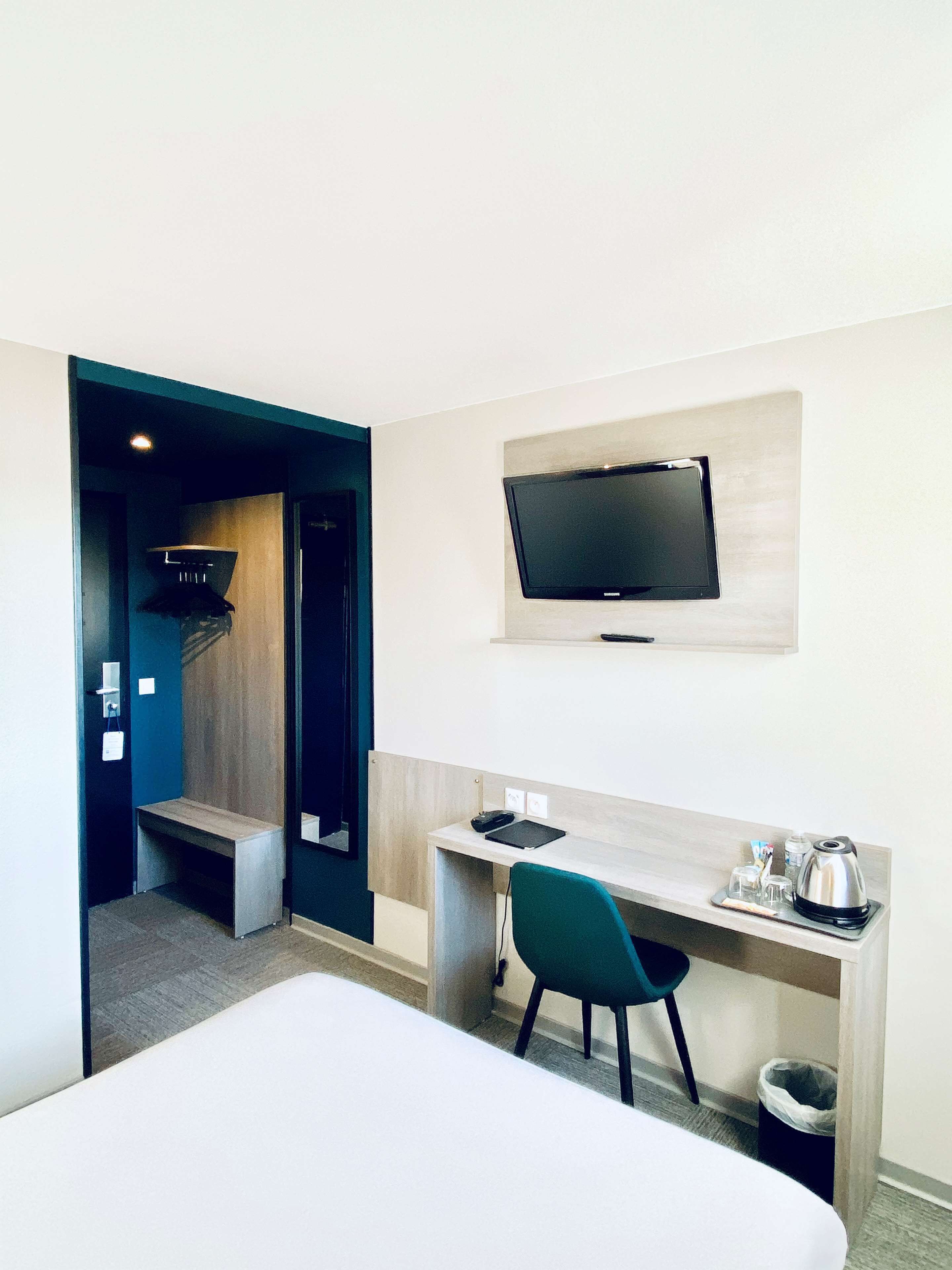 A hotel room with a bed, a desk with a chair, a TV mounted on the wall, and a small trash bin.