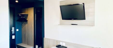 A hotel room with a bed, a desk with a chair, a TV mounted on the wall, and a small trash bin.