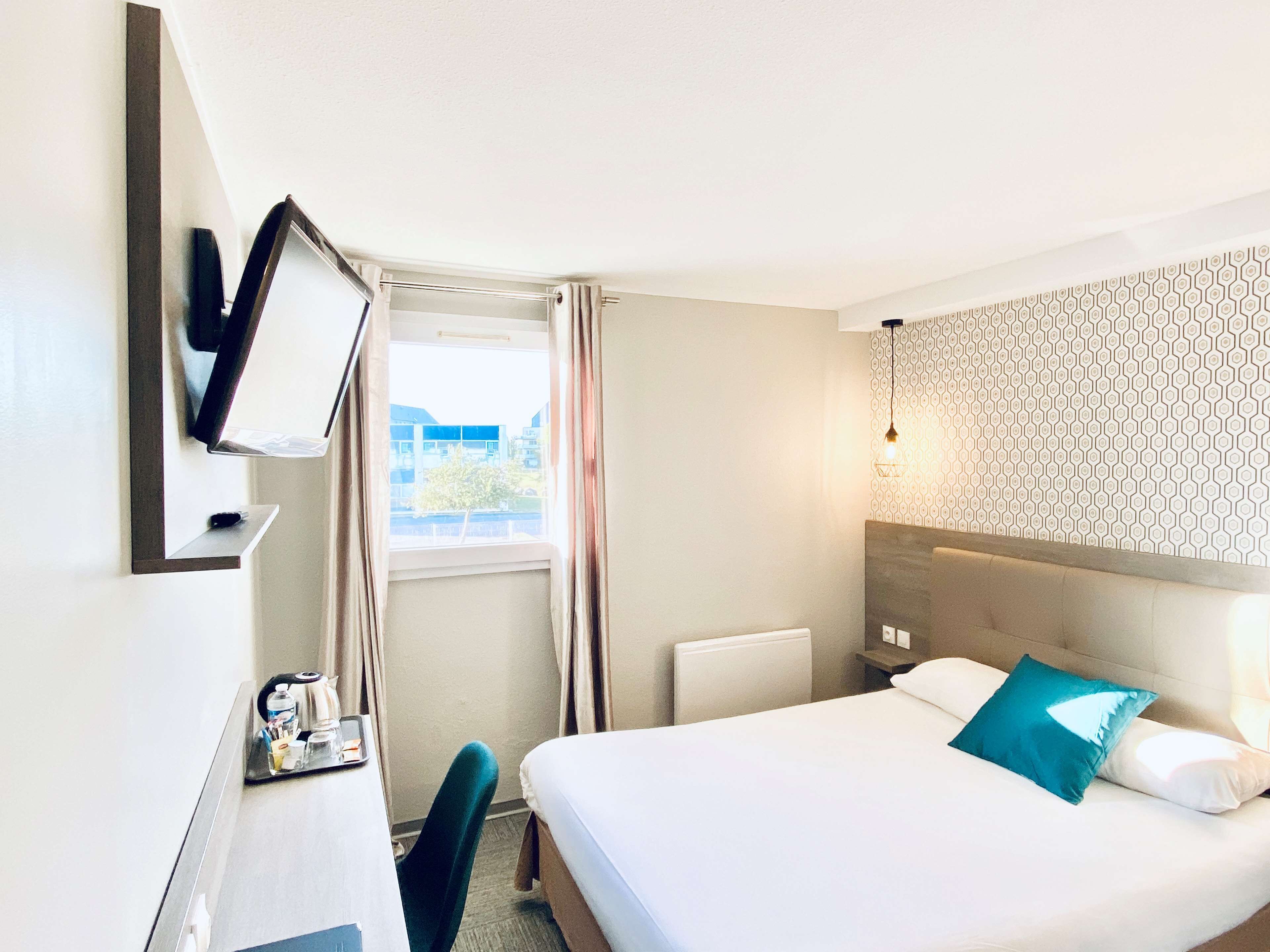 Habitación de hotel con cama, escritorio con silla, televisor montado en la pared y ventana con cortinas.