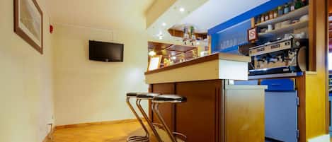 A kitchen area with a bar stool, a television mounted on the wall, and a shelf with various kitchen items.