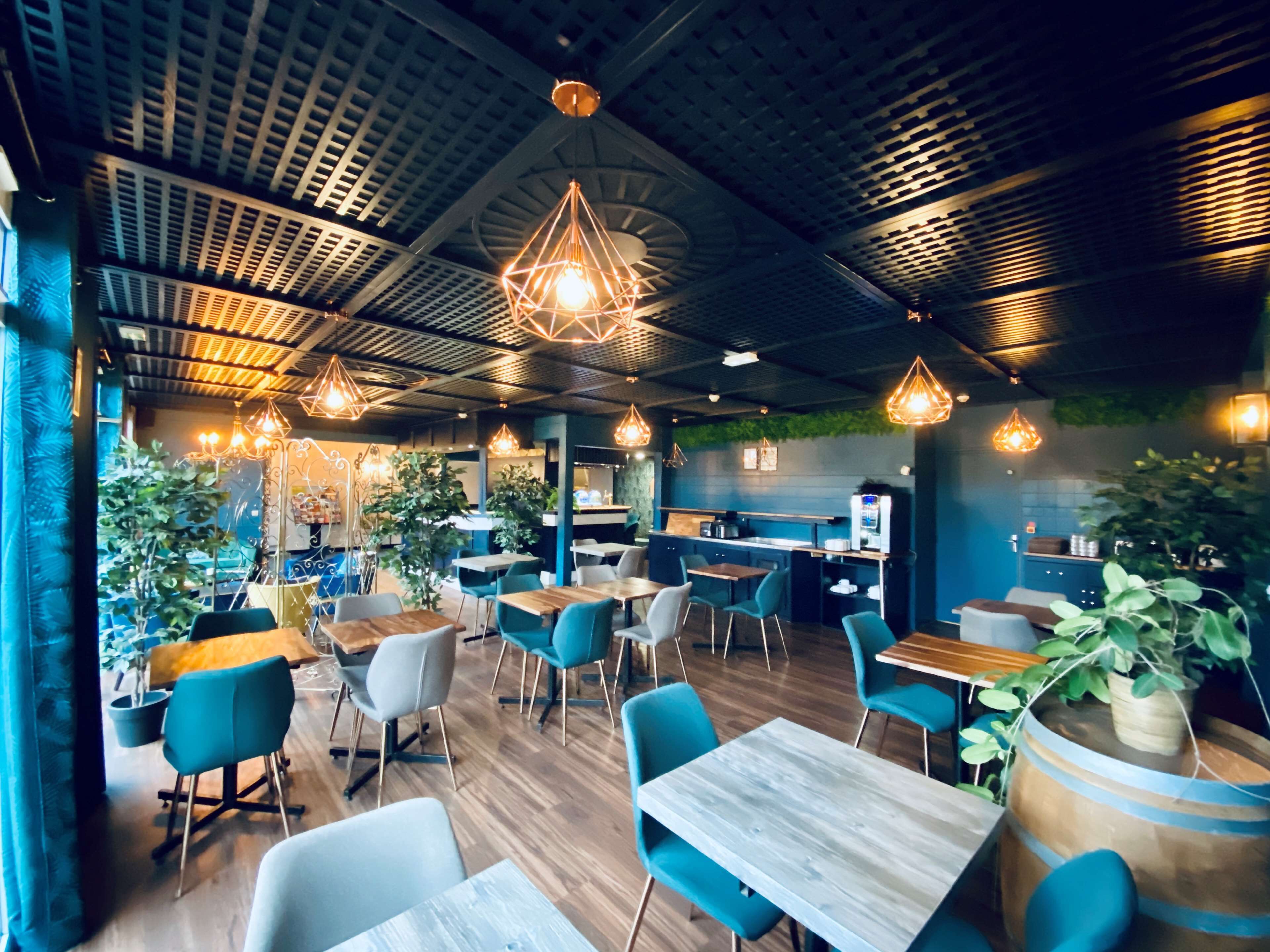 A modern restaurant interior with wooden tables and blue chairs, geometric pendant lighting, and a kitchen area in the background.