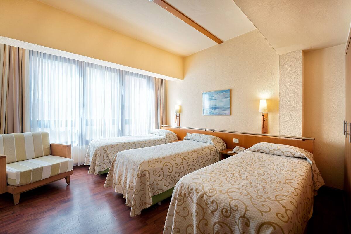 A hotel room with two beds, a sofa, a desk, and a large window with curtains.