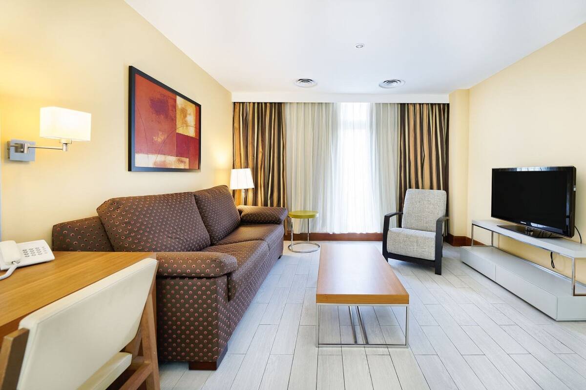 A hotel room with a sofa, a TV, a desk with a phone, a lamp, and a painting on the wall.