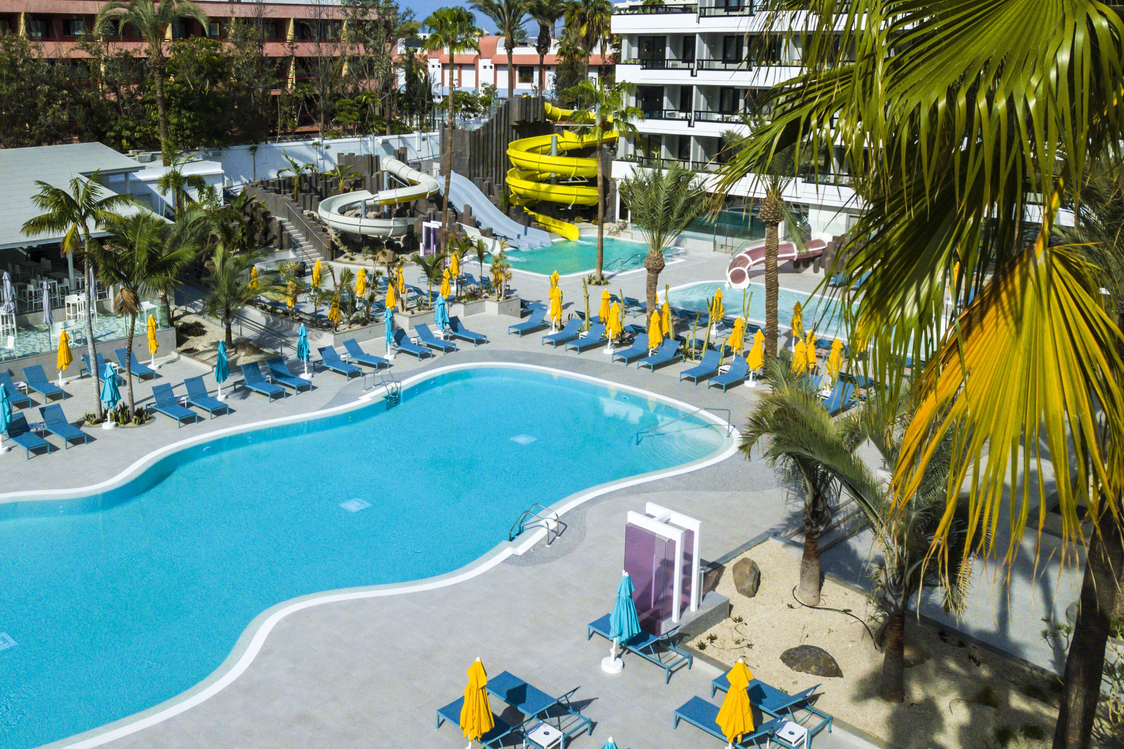 Zona de piscina en un resort con varias toboganes de agua, tumbonas y palmeras.