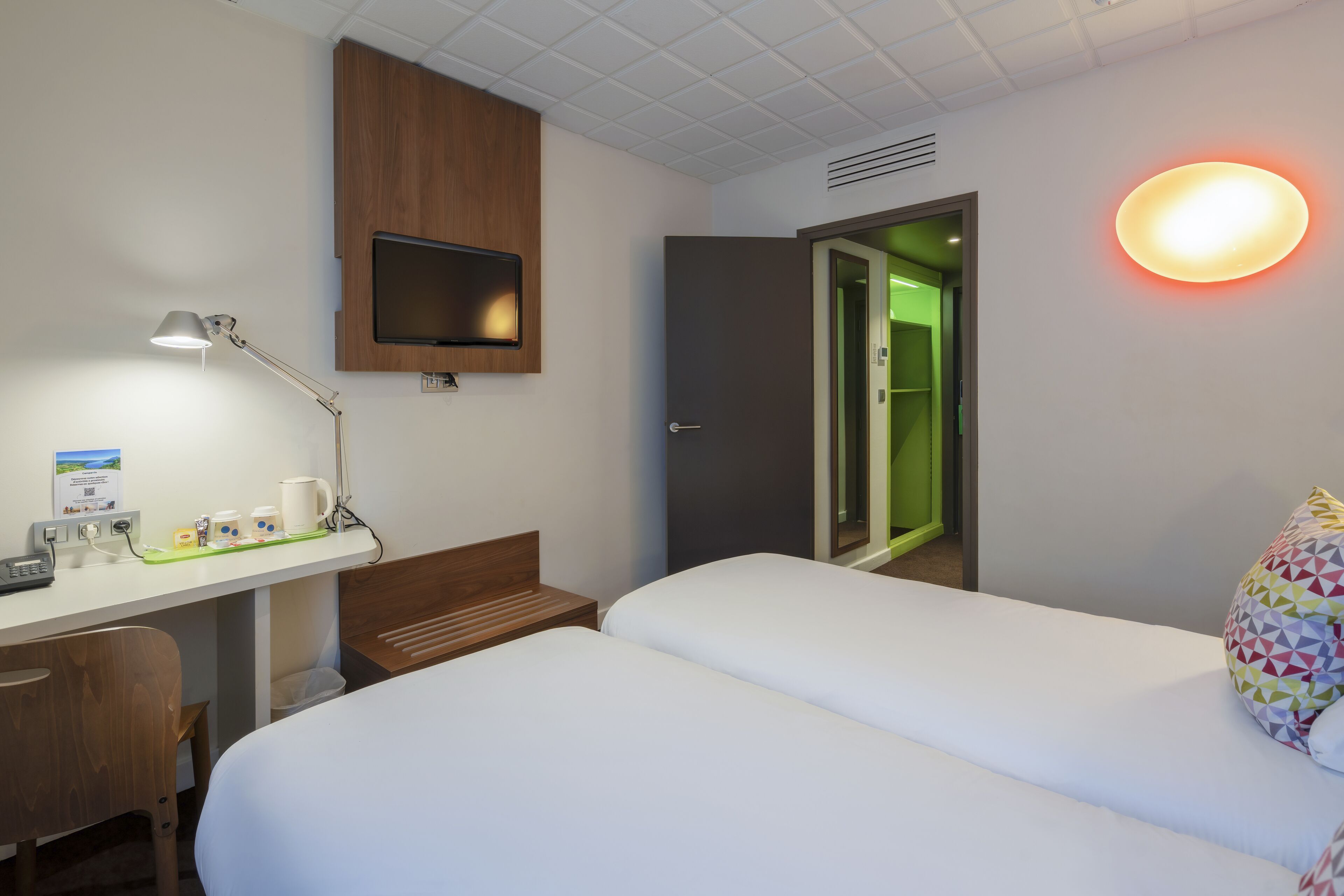 A hotel room with a desk, two beds, a TV, a lamp, and a colorful pillow.