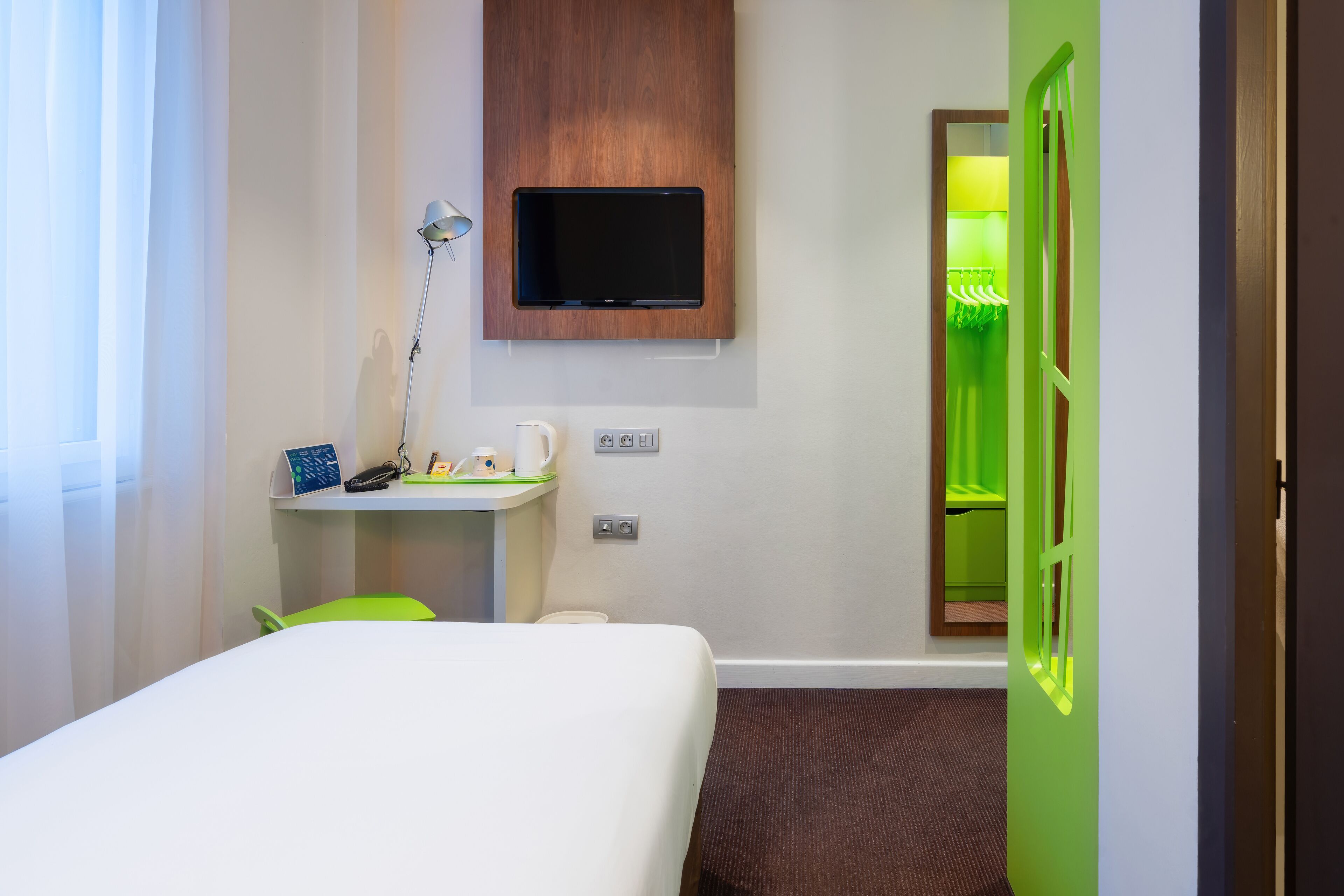 Een hotelkamer met een bed, een bureau met een lamp, een televisie en een groene deur.