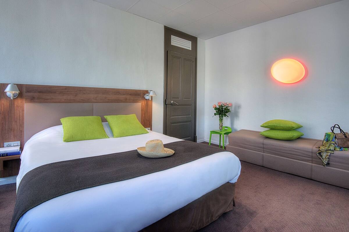 Habitación de hotel con una cama grande, cabecera de madera, mesita de noche con lámpara, almohada verde, planta en la mesita de noche y una luz en la pared.
